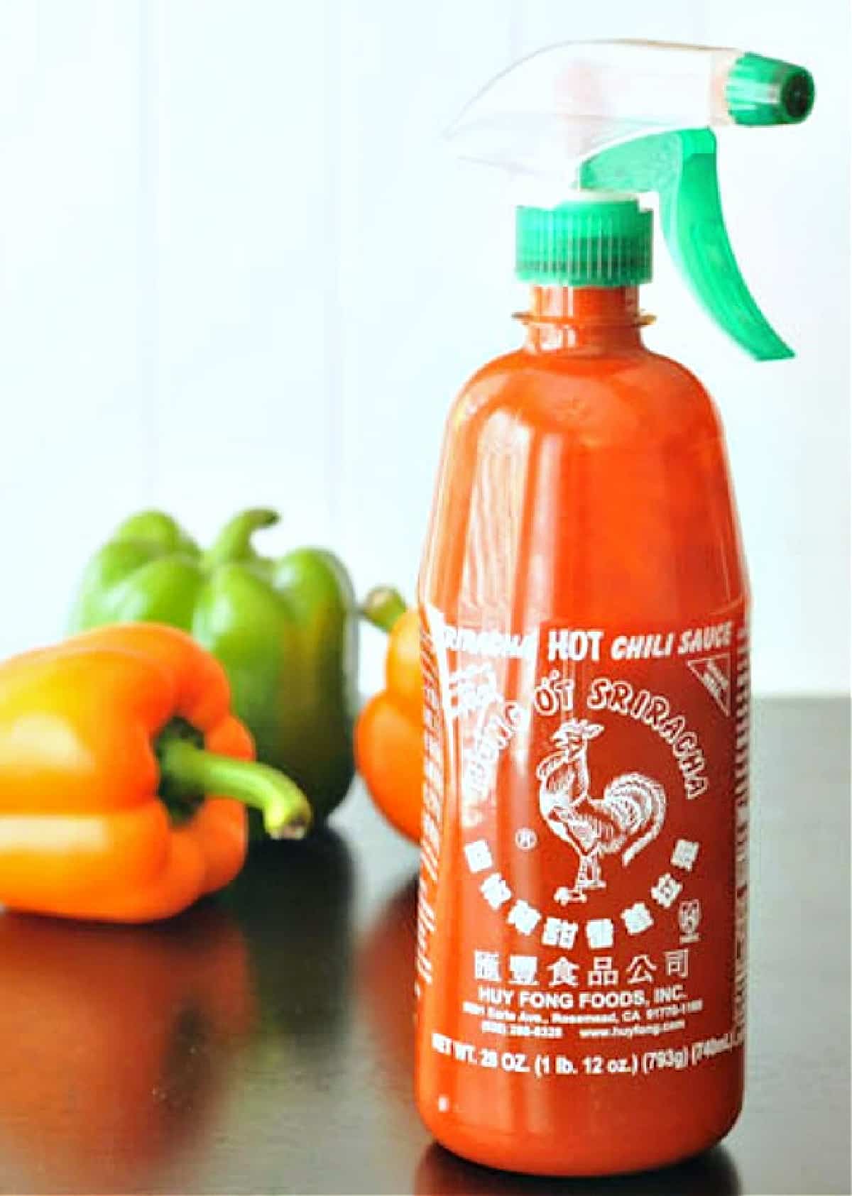 A bottle of sriracha with the normal top replaced with a nozzle sprayer. Three peppers sit behind the bottle on a black shiny surface.