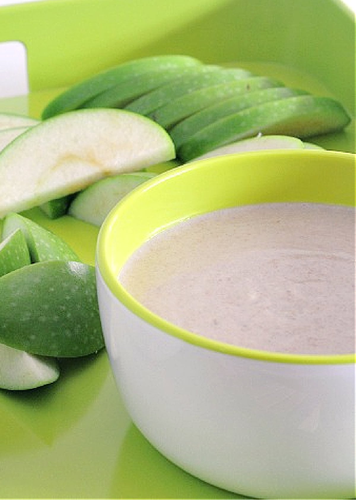 A white bowl of sweet white bean dip sits on a bright green tray with slices of tart green apple.