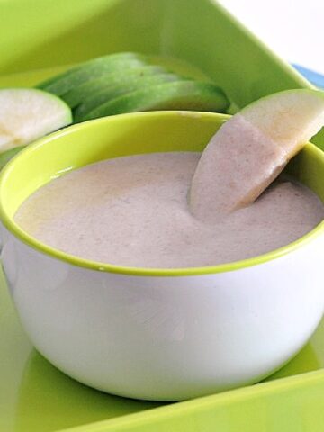 A white bowl of sweet white bean dip sits on a bright green tray with slices of tart green apple. One apple slice is dipped into the dip.