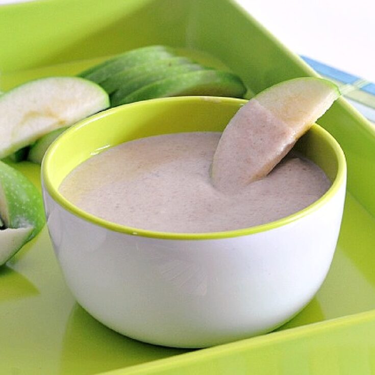 A white bowl of sweet white bean dip sits on a bright green tray with slices of tart green apple. One apple slice is dipped into the dip.