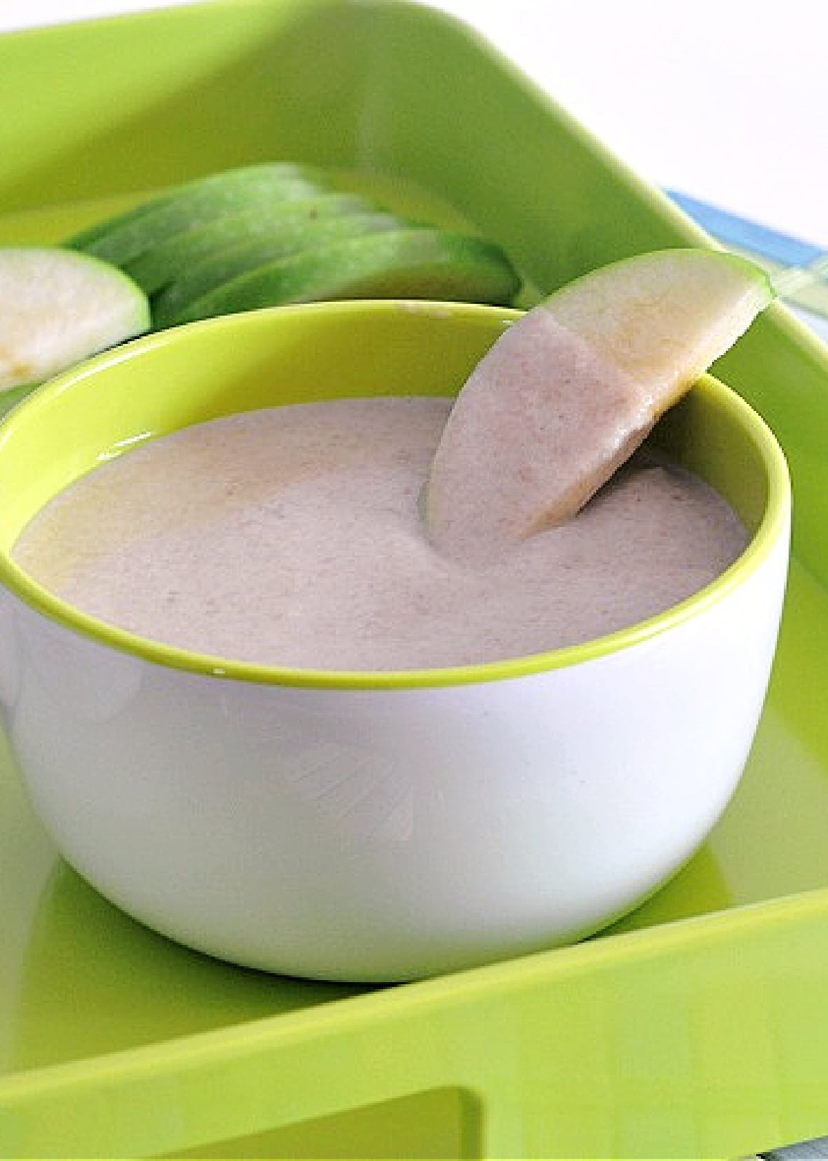 A white bowl of sweet white bean dip sits on a bright green tray with slices of tart green apple. One apple slice is dipped into the dip.