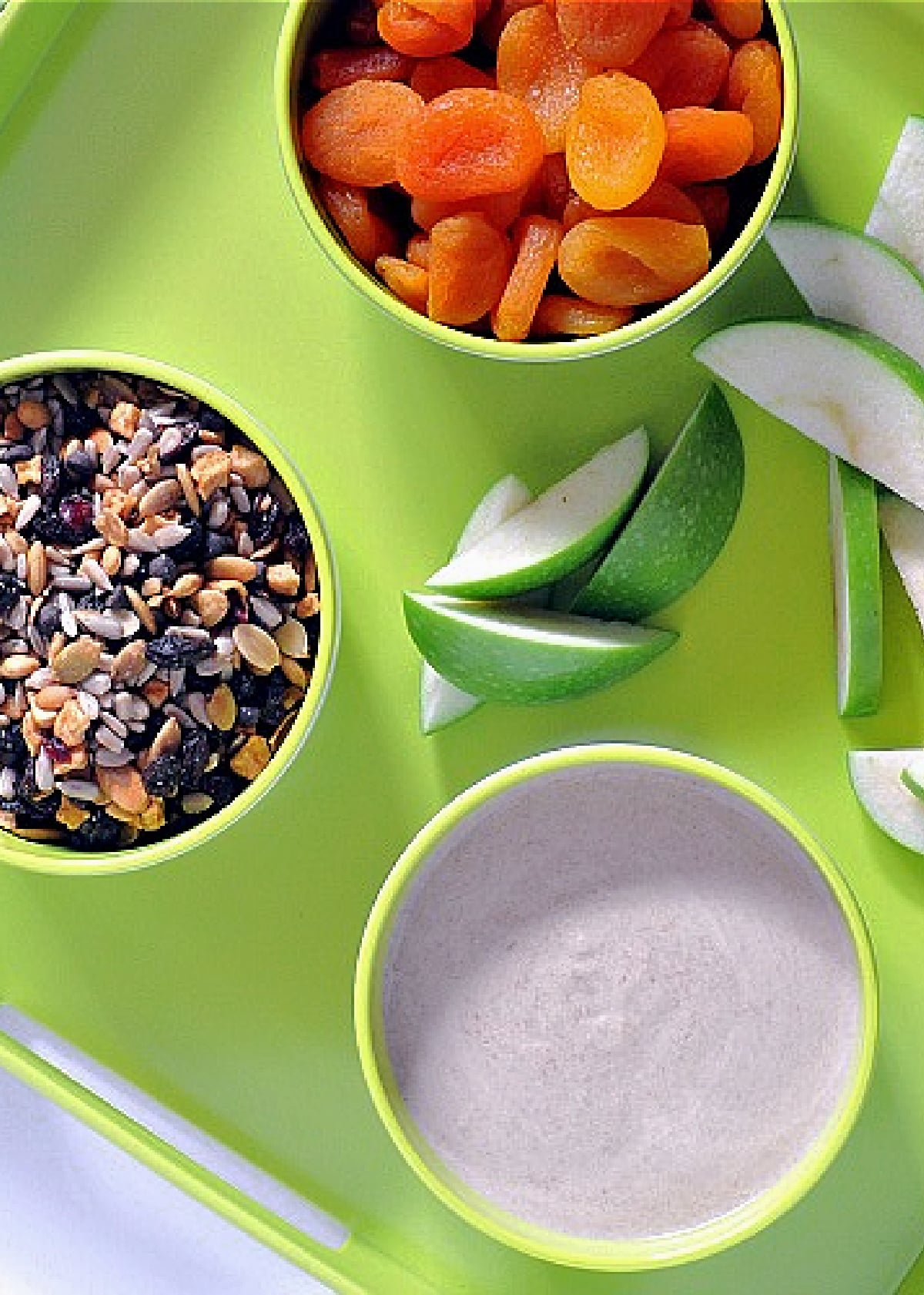 Overhead view of three white bowls on a bright green serving tray. One bowl is filled with dried apricots, one bowl is sweet white bean dip, and one bowl holds trail mix. Slices of green apple sit on the tray between the bowls.