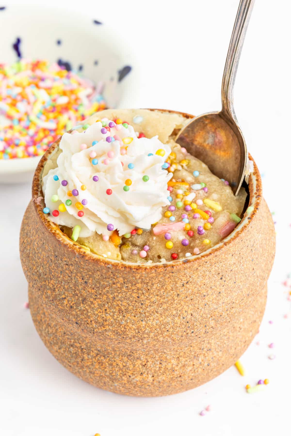 A spoon sits in a vanilla mug cake in the light brown pottery mug it was cooked in. Cake is yellow with sprinkles and is topped with whipped cream.