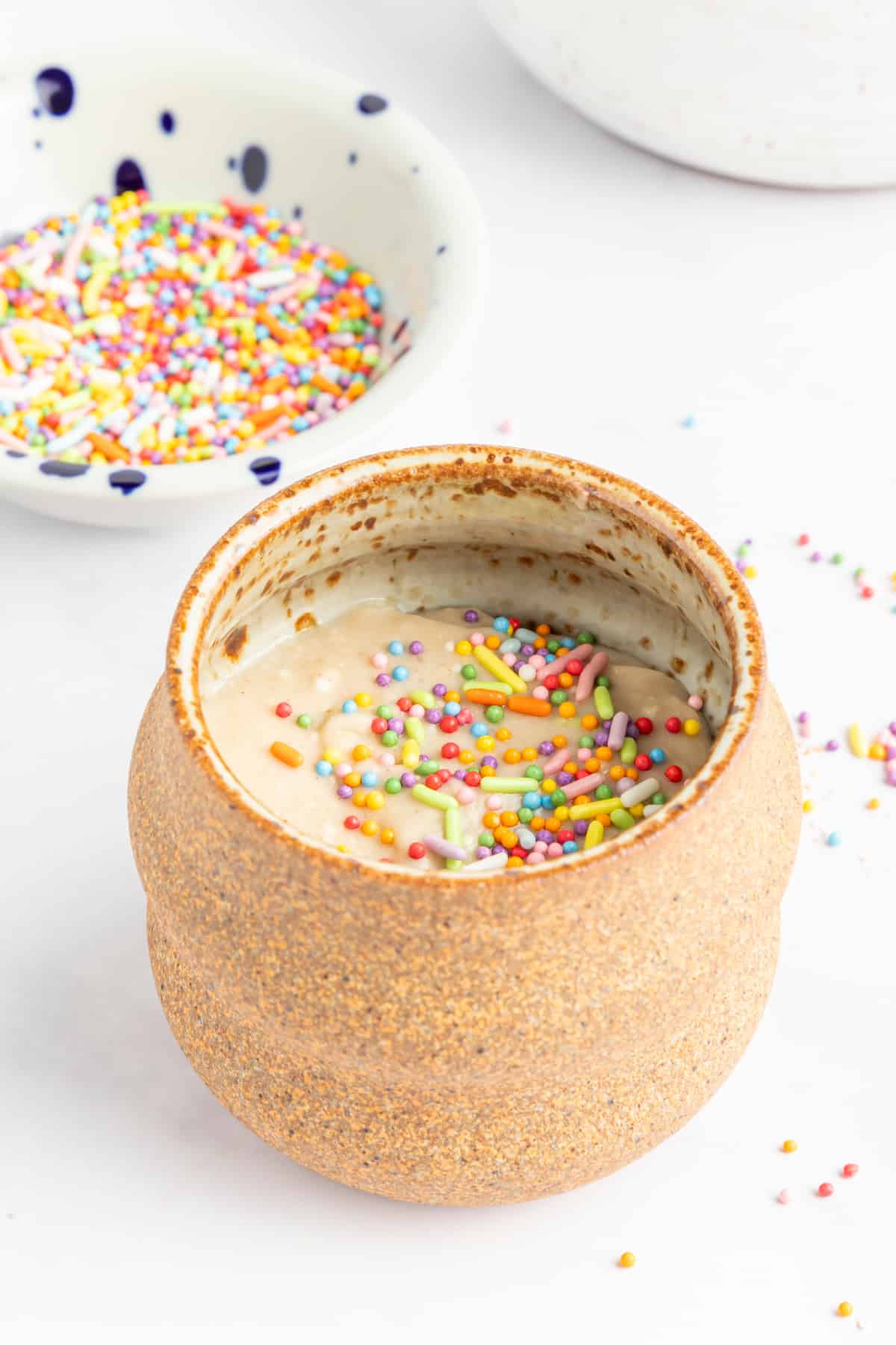 The batter for a minute mug muffin sits in the mug before cooking. Mug is light brown pottery, sitting on a white background next to a small bowl of pastel colored sprinkles.
