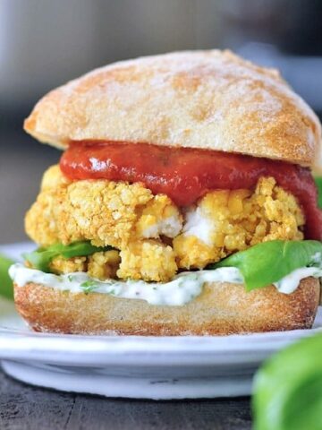 Close up view of a vegan mozzarella chicken burger in a bun with basil aioli and marinara sauce sits on a white plate. A basil plant is blurred in the foreground.