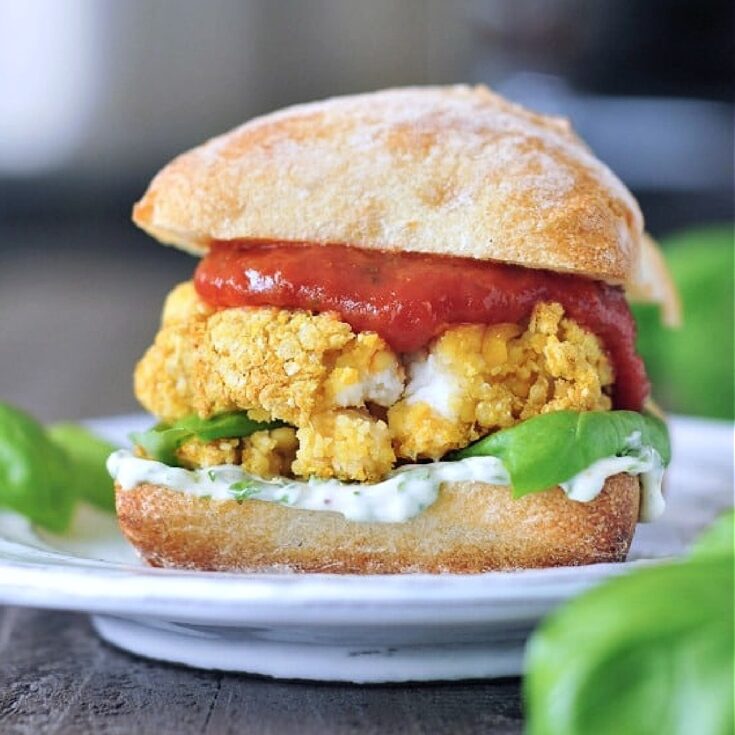 Close up view of a vegan mozzarella chicken burger in a bun with basil aioli and marinara sauce sits on a white plate. A basil plant is blurred in the foreground.