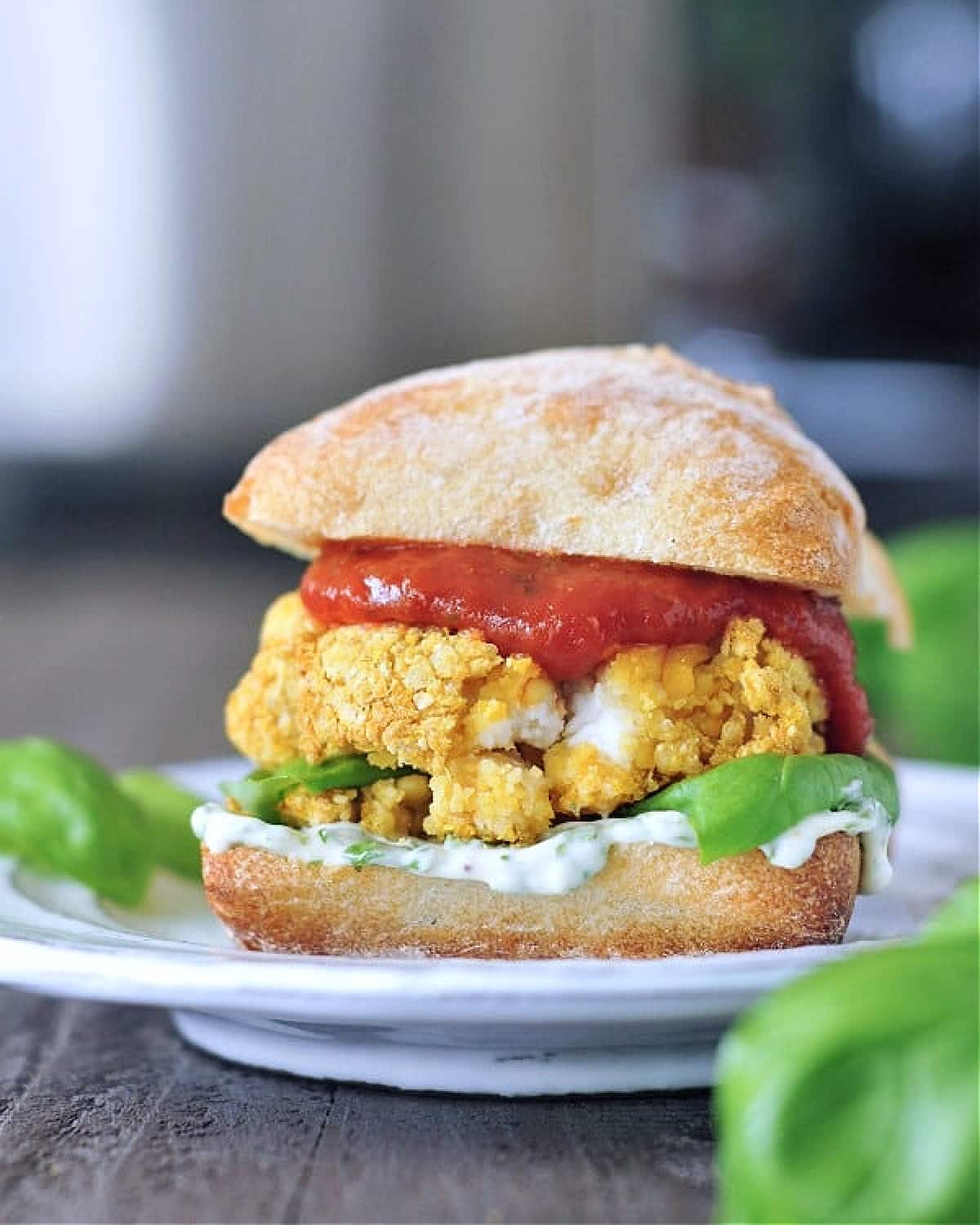 A vegan mozzarella stuffed chicken burger in a bun with basil aioli and marinara sauce sits on a white plate. A basil plant is blurred in the foreground.
