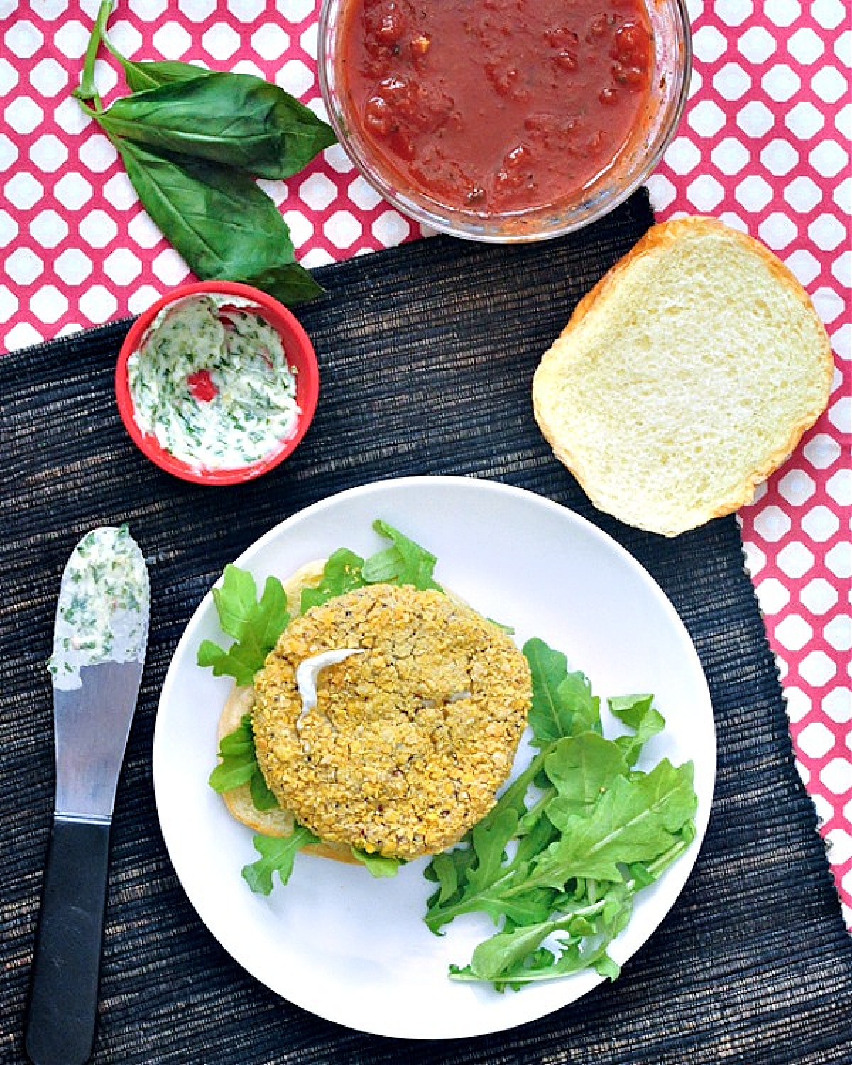 Overhead view of a vegan chicken patty on a bottom bun with arugula, sitting on a white plate. The top bun sits to the side with a basil aioli, a knife with aioli on it, and a small glass bowl of marinara sauce.