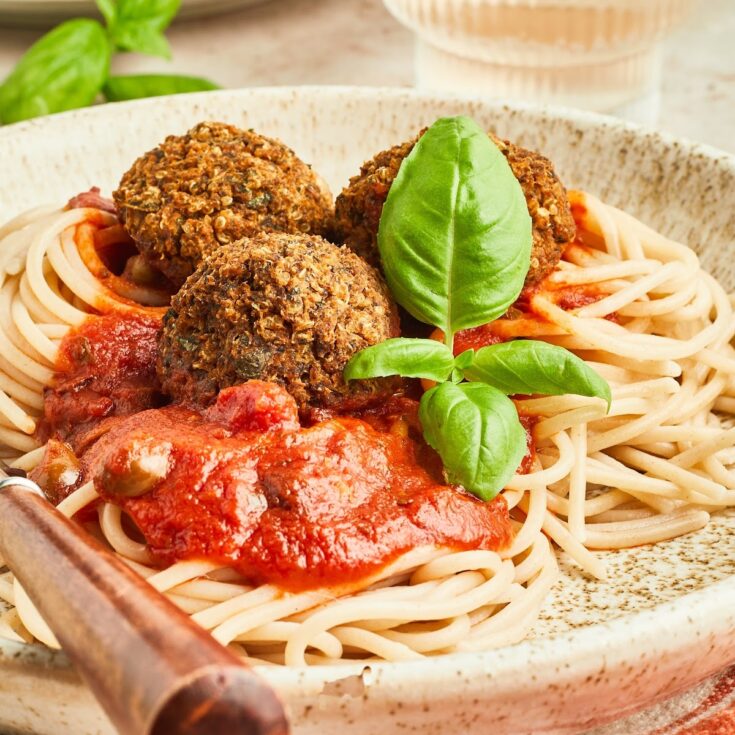 Close up of three vegetarian meatballs over spaghetti noodles with marinara sauce on a light beige rustic plate with a wood handled fork. Food is garnished with fresh basil, and the plate sits on a red and white striped cloth napkin.