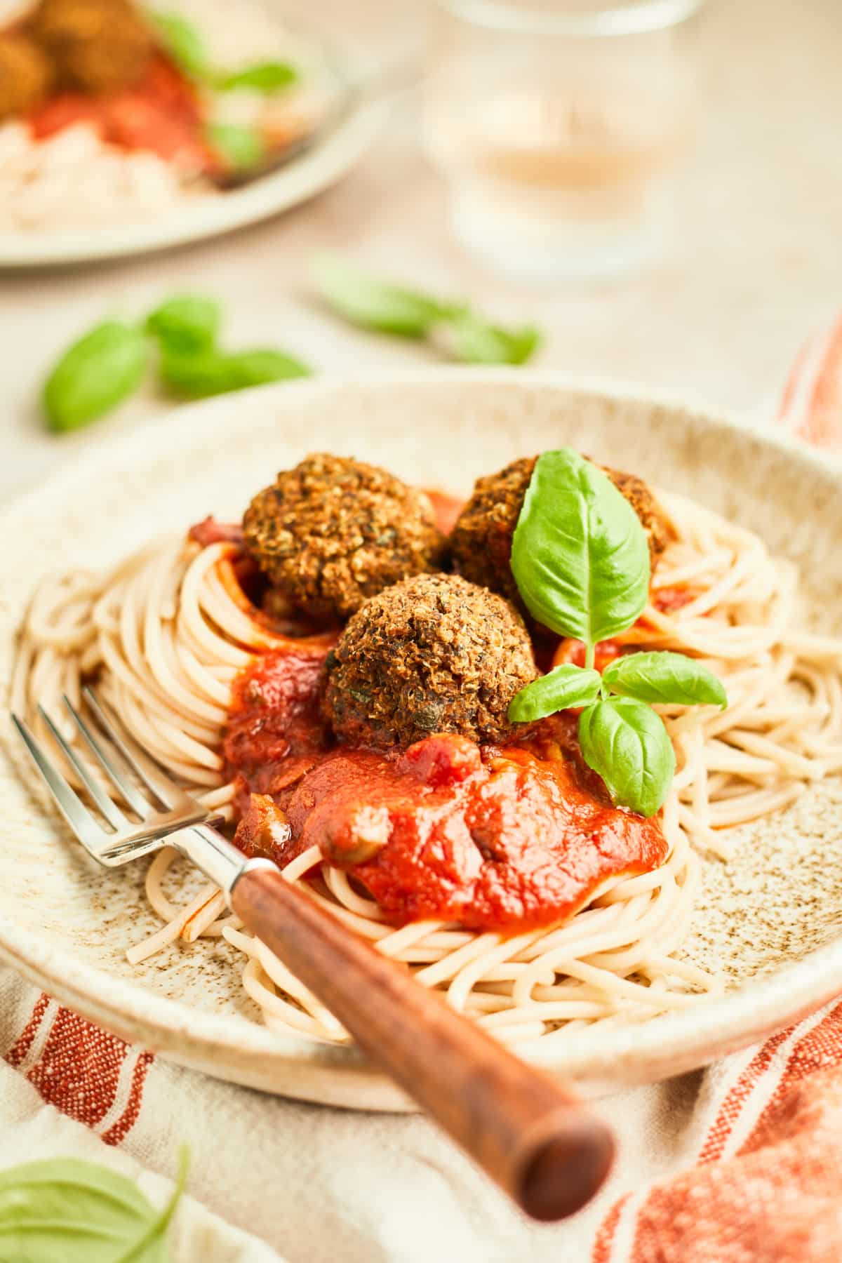 Three basil quinoa meatballs over spaghetti noodles with marinara sauce on a light beige rustic plate with a wood handled fork. Food is garnished with fresh basil, and the plate sits on a red and white striped cloth napkin.