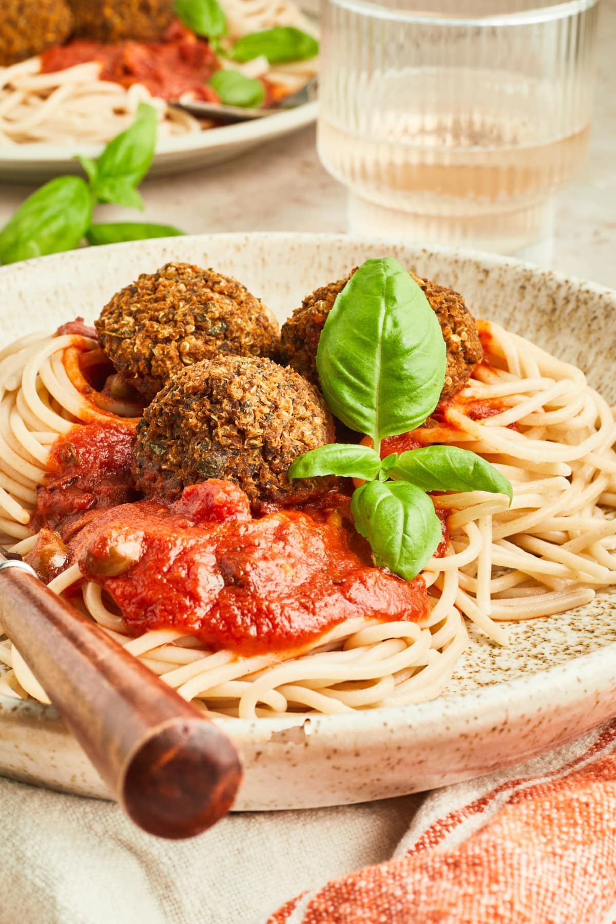 Three basil quinoa meatballs over spaghetti noodles with marinara sauce on a light beige rustic plate with a wood handled fork. Food is garnished with fresh basil, and the plate sits on a red and white striped cloth napkin. Another similar plate of food sits off focus in background with a glass of white vwine.