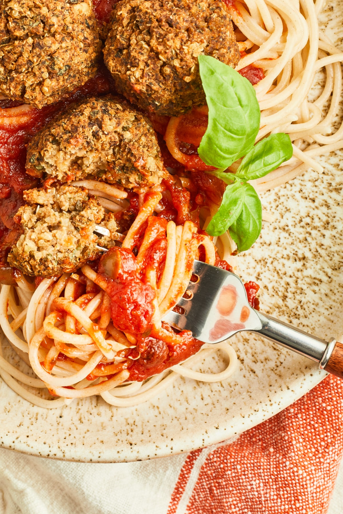 Overhead view of basil quinoa meatballs on spaghetti and marinara sauce. A fork has spaghetti noodles wrapped around it, and one meatball is sliced in half to show texture inside. Fresh basil garnishes the plate.