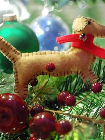 A handmade felt Christmas ornament sewn in the shape of a dachshund dog with a red scarf around the neck. Dachshund ornament is on a holiday tree with other ornaments and berries.