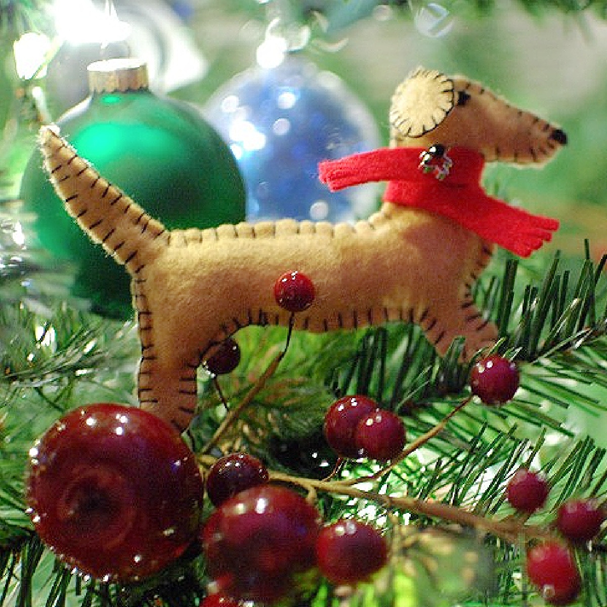 A handmade felt Christmas ornament sewn in the shape of a dachshund dog with a red scarf around the neck. Dachshund ornament is on a holiday tree with other ornaments and berries.