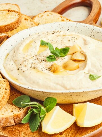 A shallow white bowl of creamy white bean dip garnished with several roasted garlic cloves and fresh oregano leaves sits on a round wooden board with fresh lemon wedges and toasted baguette slices.