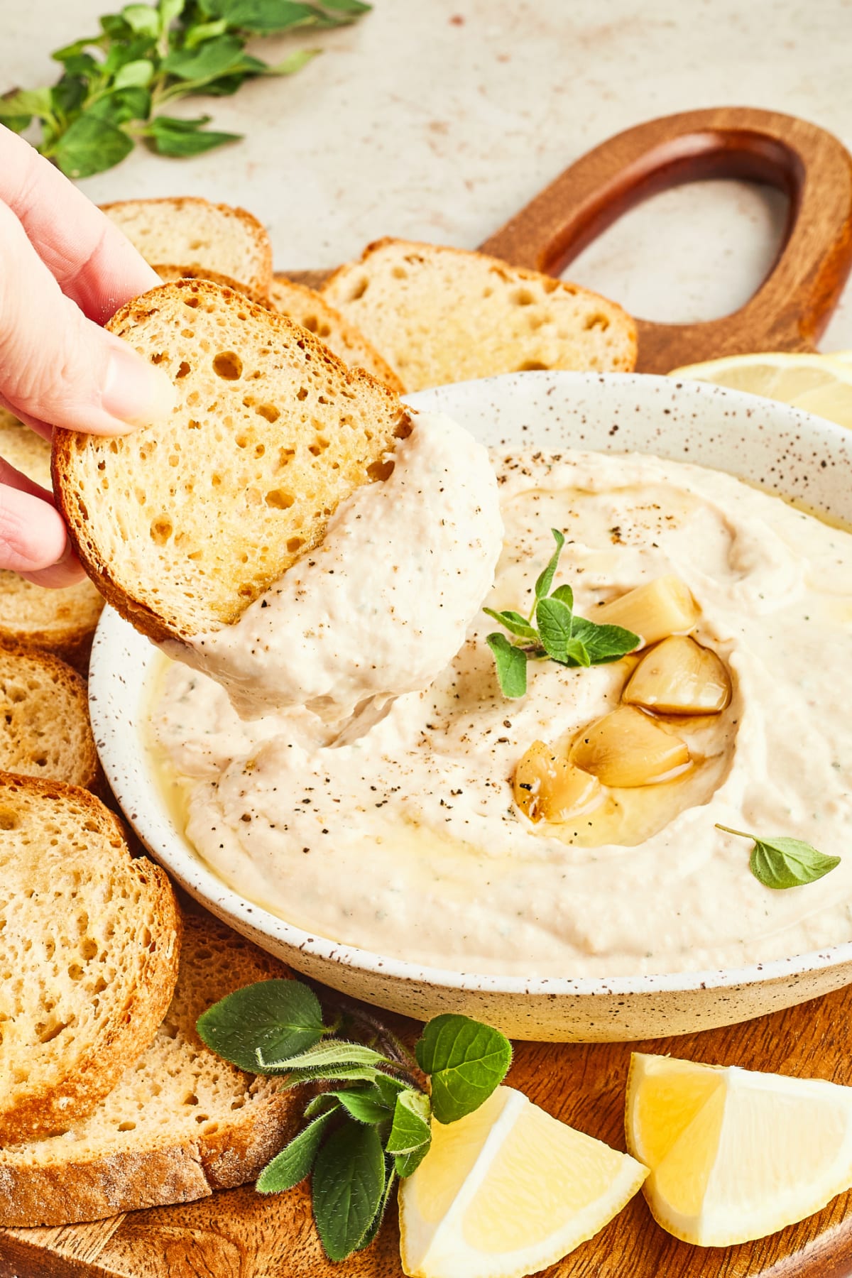 A hand dipping a slice of toasted baguette into a bowl of white bean dip. The bowl of dip is garnished with several roasted garlic cloves and fresh oregano leaves, and sits on a round wooden board with fresh lemon wedges and toasted baguette slices.