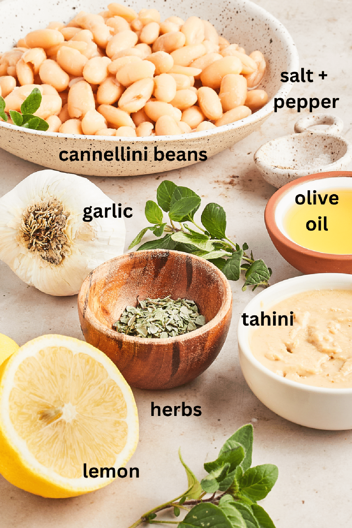 Several bowls of ingredients to make roasted garlic dip: cannelini beans, spices, herbs, garlic, tahini, olive oil, and fresh lemon.