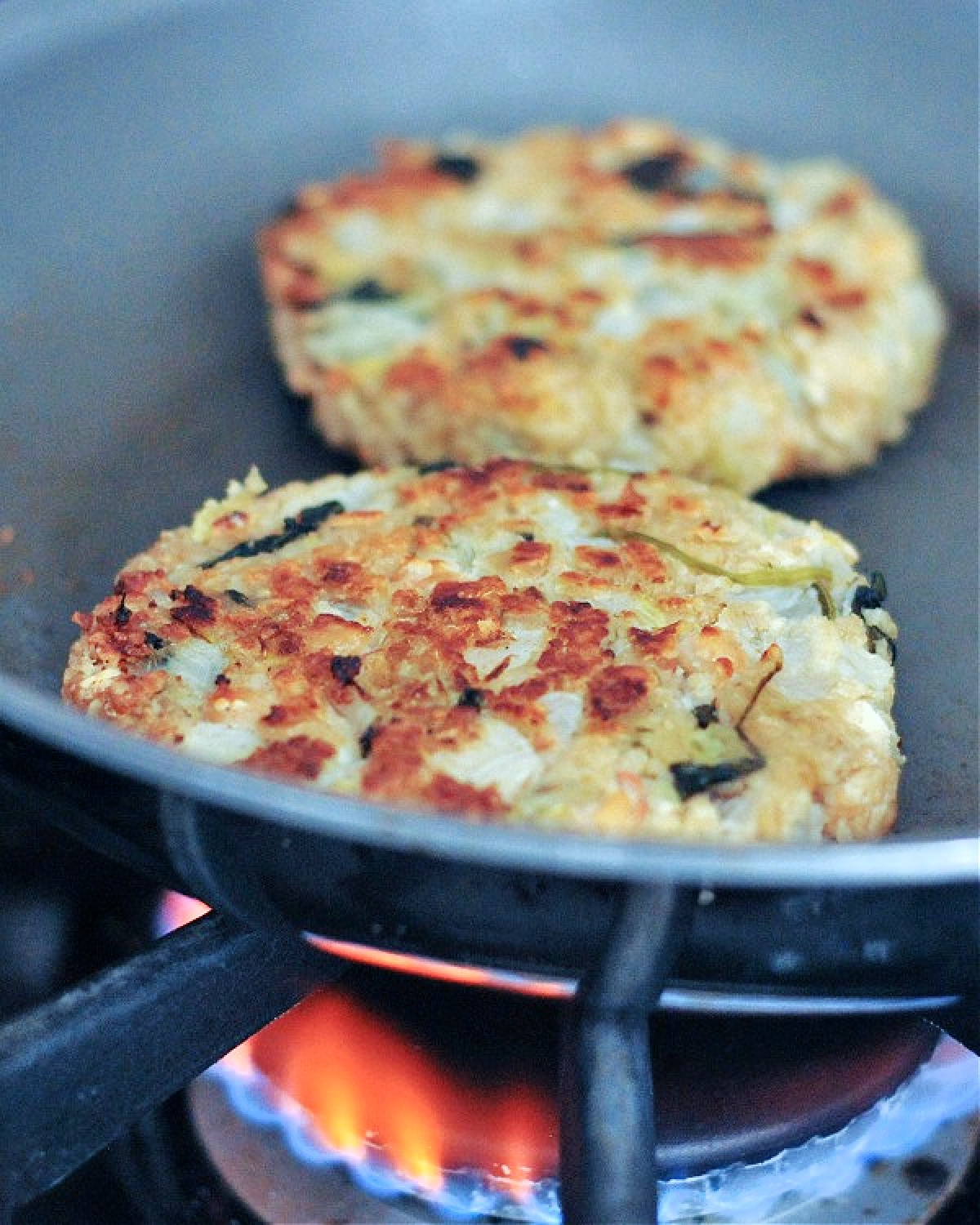 Two spinach artichoke burger patties sit cooking in a skillet over a gas fire cooktop.