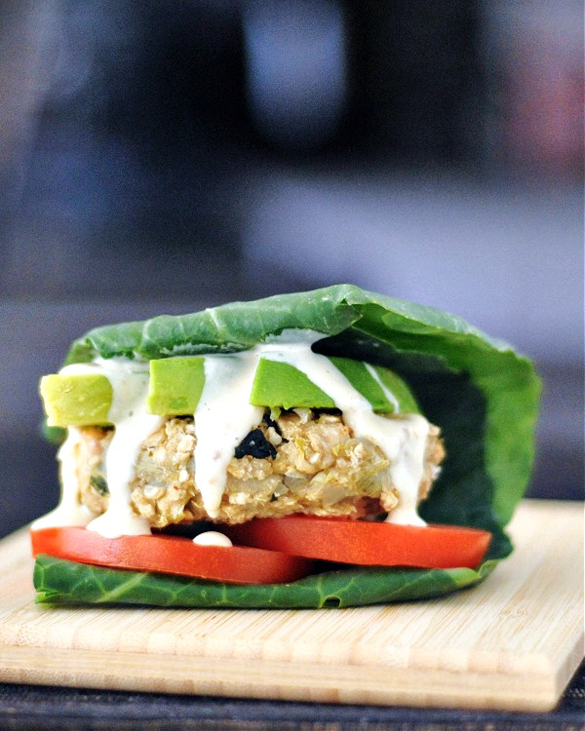 A veggie burger is topped with tomato, avocado, and wrapped in a fresh collard leaf. Burger sits on a small wooden serving board.
