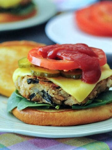 A veggie burger topped with cheese, pickles, tomato slices, and ketchup sits on a light green plate. Another plate of pickles and tomato slices is blurred in background, and the burger plate sits on a pastel plaid cloth napkin.