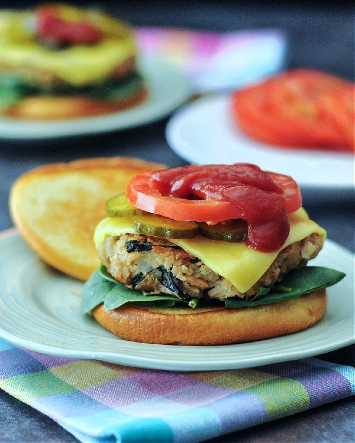A veggie burger topped with cheese, pickles, tomato slices, and ketchup sits on a light green plate. Another plate of pickles and tomato slices is blurred in background, and the burger plate sits on a pastel plaid cloth napkin.