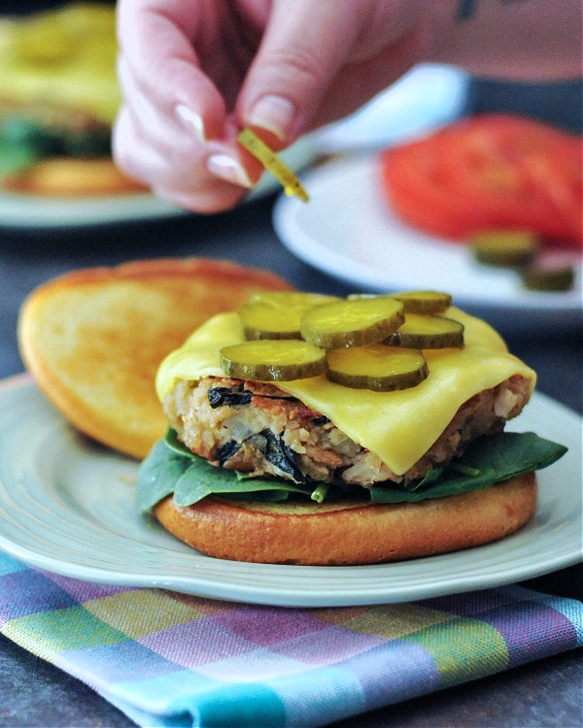 A hand adding pickle chips to the top of a cheeseburger sitting on a light green plate. Another plate of pickles and tomato slices is blurred in background, and the burger plate sits on a pastel plaid cloth napkin.