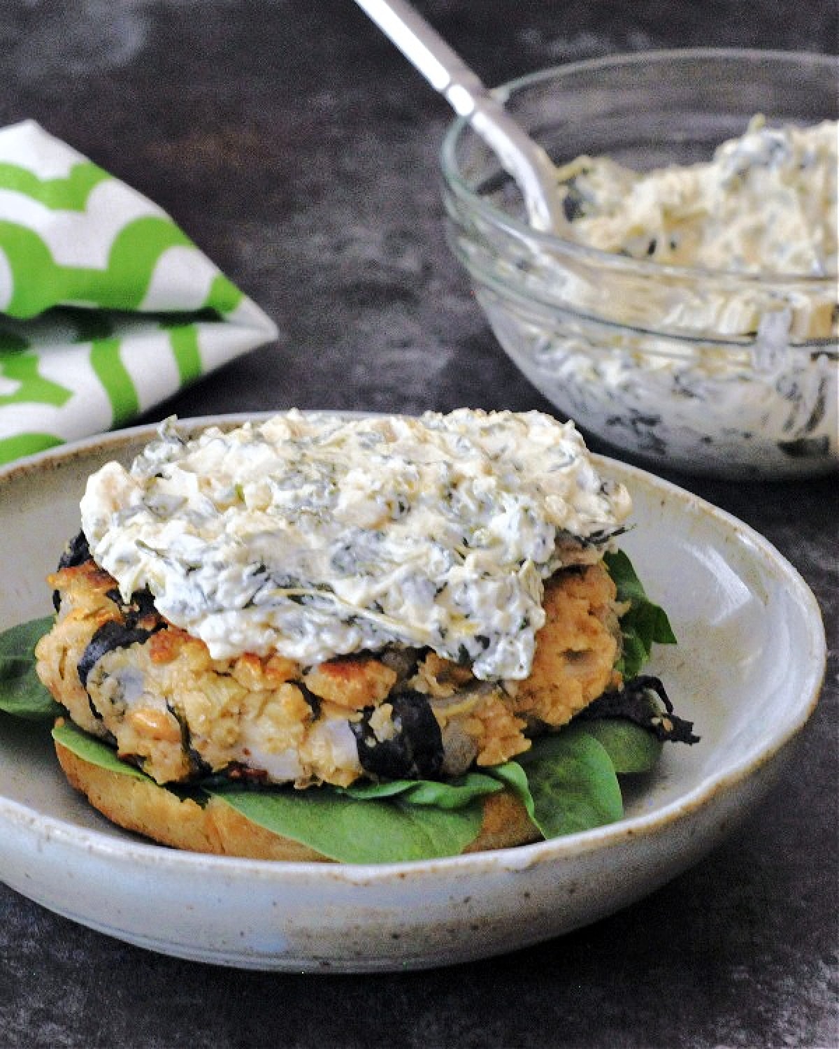 A veggie burger sits on a bun with fresh spinach. The burger is topped with spinach dip, and there is a small glass bowl of spinach dip next to the plate with the burger.