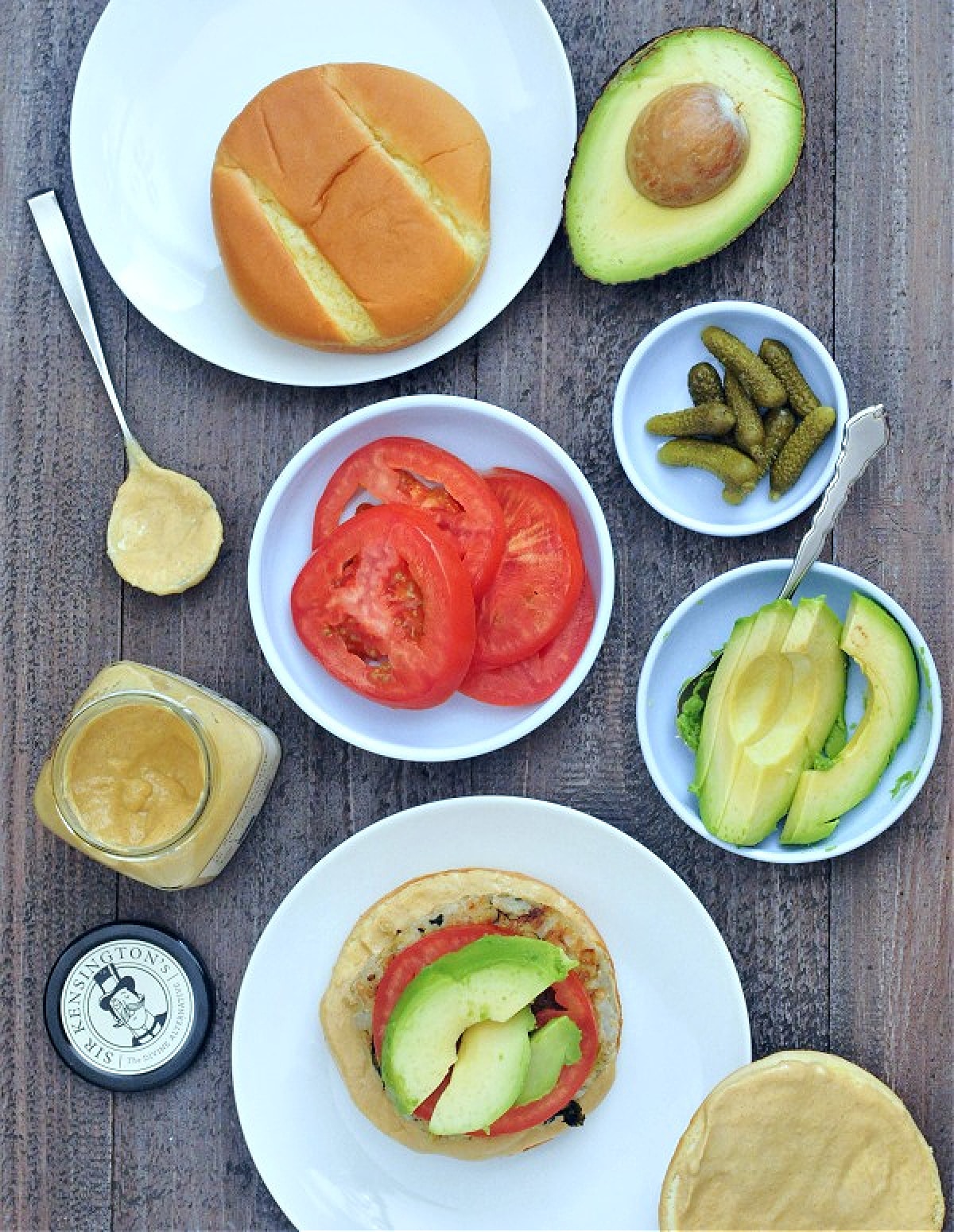 Overhead view of a burger on a bottom bun, sitting on a white plate. Burger is dressed with a tomato slice and three slices of avocado. Next to this plate, there is an open jar of mustard and a mustard covered spoon, a half avocado and a small plate of avocado slices, a small plate of tomato slices and another plate with gherkin pickles.