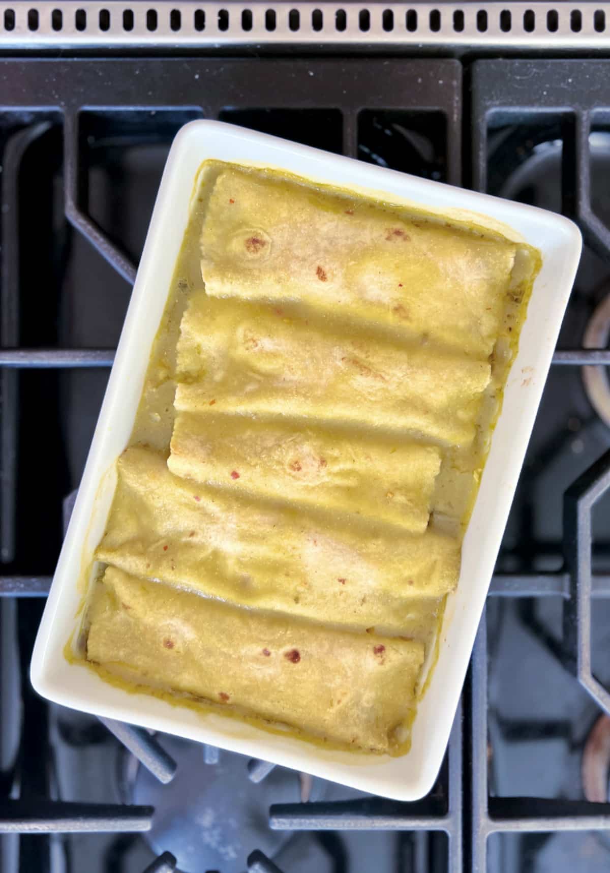 Overhead view of a white ceramic baking dish of breakfast enchiladas sitting on a cooktop. Five enchiladas lined up and covered with green enchilada sauce, fresh baked just out of the oven.