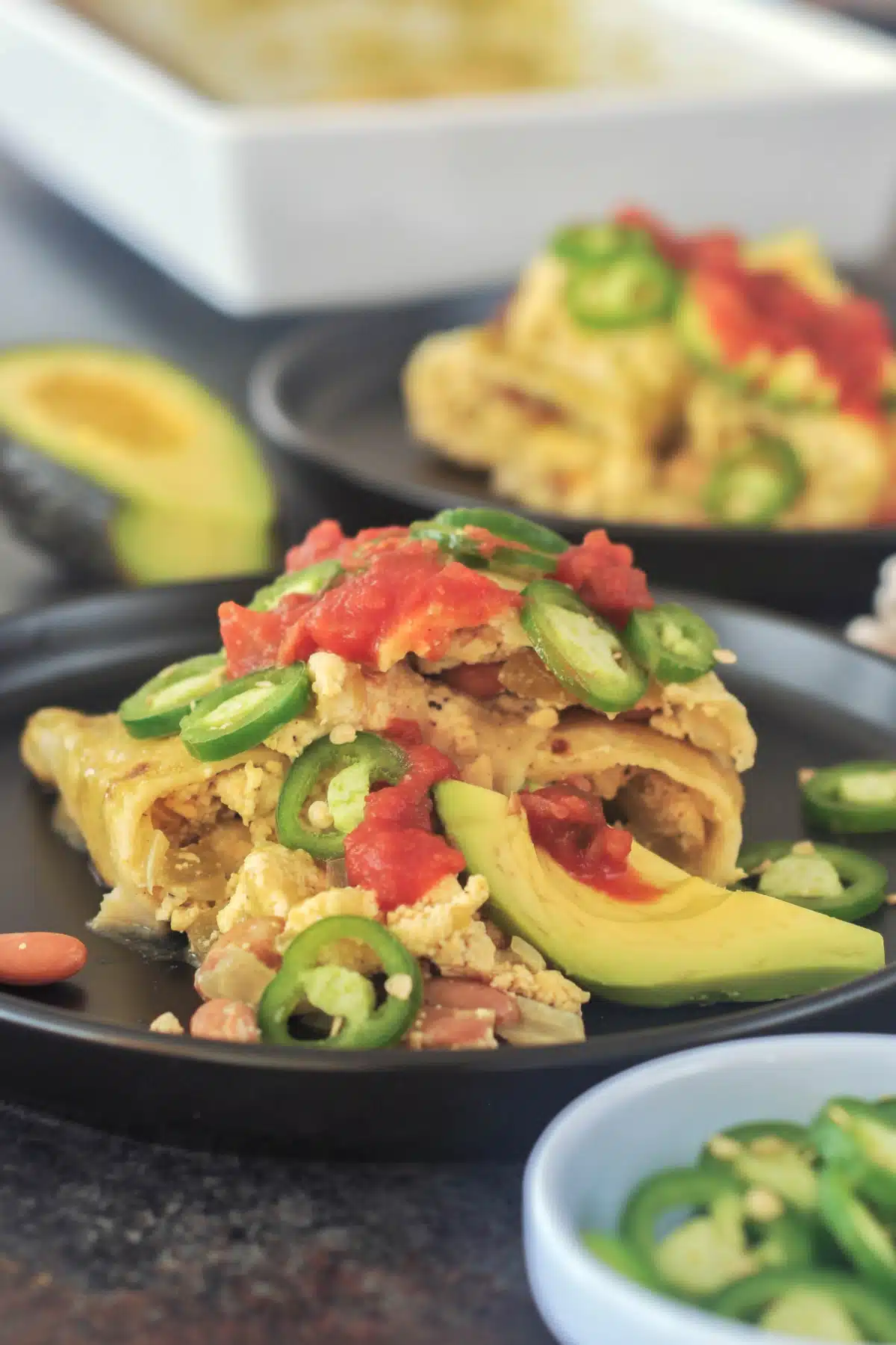 One serving of breakfast enchiladas on a matte black plate. Enchiladas are filled with a tofu scramble and beans, and garnished with sliced jalapeños, sliced avocado, and deep red salsa. A half avocado and the baking dish of enchiladas are blurred in the background.