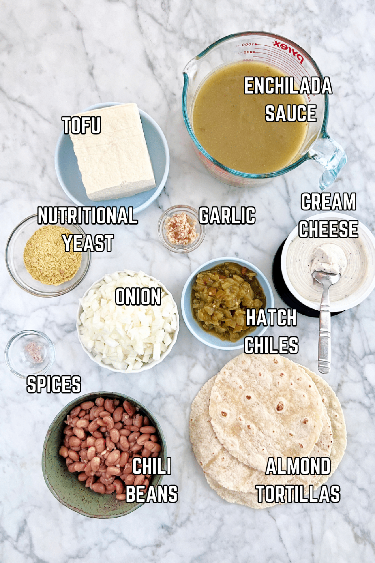 Overhead view of several small bowls of ingredients to make vegan breakfast enchiladas sit on a white marble surface: green enchilada sauce, tofu, nutritional yeast, garlic, onion, spices, Hatch chiles, chili beans, cream cheese, and tortillas.