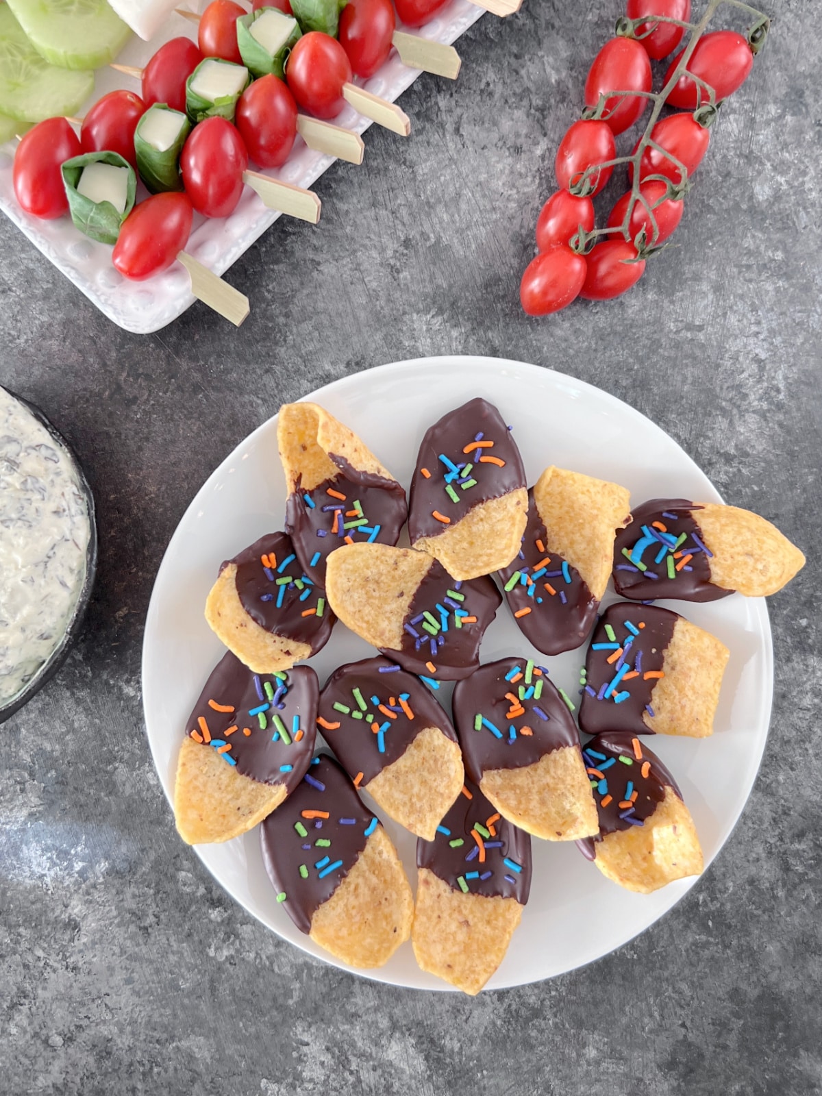 Overhead view of a white plate of chocolate dipped corn chips with multi colored sprinkles in the chocolate. Another serving plate of veggies and crackers is blurred in background on a table set for a party.