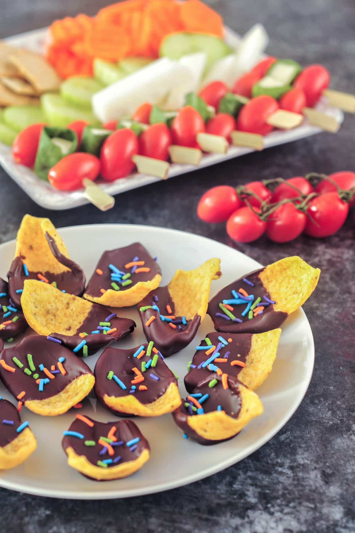A white plate of chocolate dipped corn chips with multi colored sprinkles in the chocolate. Another serving plate of veggies and crackers is blurred in background on a table set for a party.