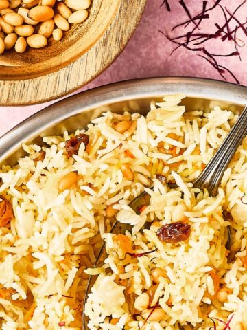 A close up overhead view of a serving bowl of yellow saffron rice garnished with golden raisins, pine nuts, a cinnamon stick and saffron strands. A small wooden bowl of toasted pine nuts sits to the side of the rice, and a serving spoon is in the bowl of rice.