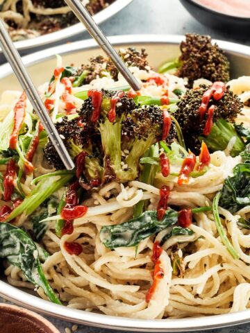 Close up of a bowl of ginger sesame noodles with roasted broccoli. A pair of chopsticks are lifting a piece of broccoli from the noodle bowl.