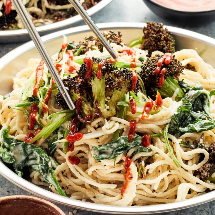 Close up of a bowl of ginger sesame noodles with roasted broccoli. A pair of chopsticks are lifting a piece of broccoli from the noodle bowl.