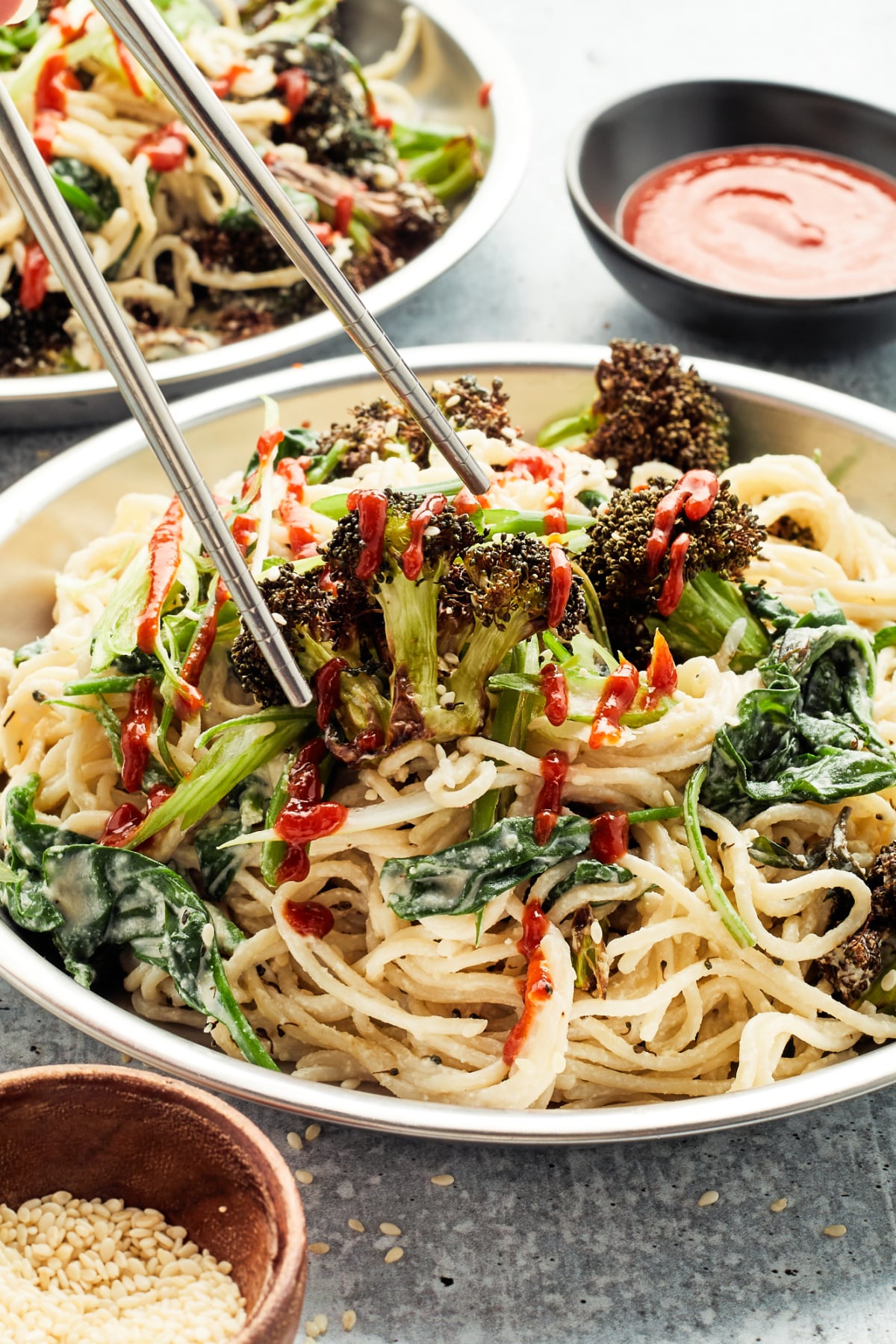 Two bowls of ginger sesame noodles with roasted broccoli. Silver chopsticks are about to lift a piece of broccoli from one bowl of noodles. A small bowl of sriracha sauce and a small bowl of sesame seeds sit to the side of the noodle bowls.
