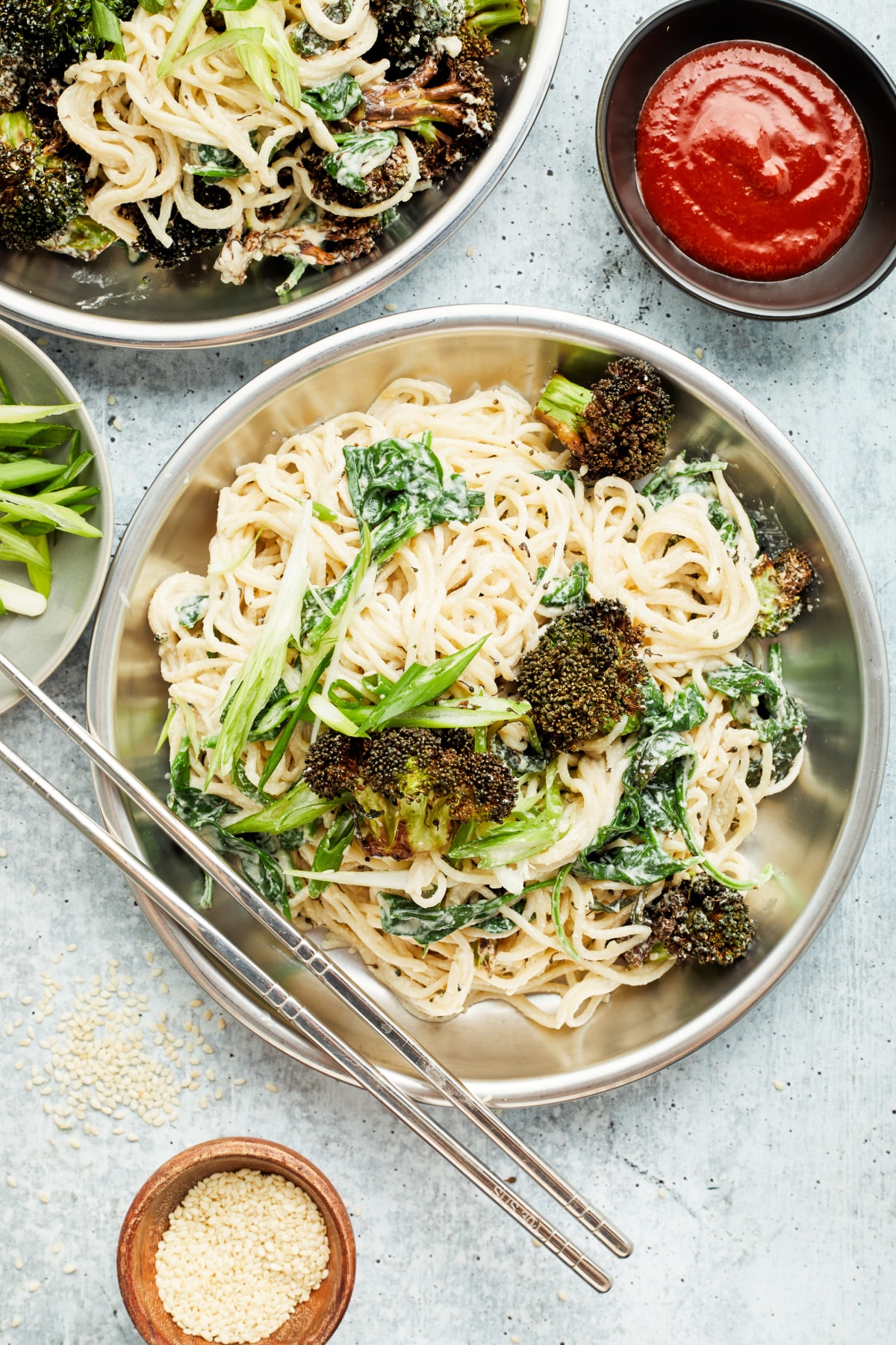 Overhead view of two bowls of ginger sesame noodles with roasted broccoli. A set of silver chopsticks lays across one bowl, and small bowls of sriracha sauce and sesame seeds sit to the side of the noodle bowls.