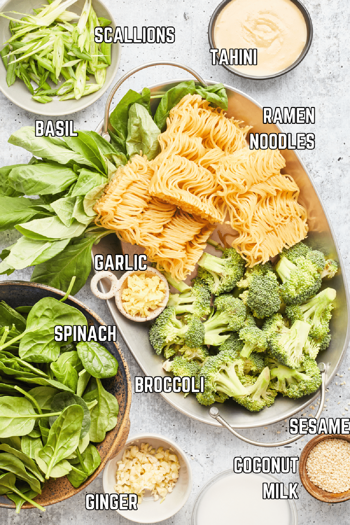 Overhead view of several bowls of ingredients to make ginger sesame noodles: scallions, tahini, fresh basil, ramen noodles, fresh minced garlic, broccoli, spinach, fresh minced ginger, sesame seeds, and coconut milk.