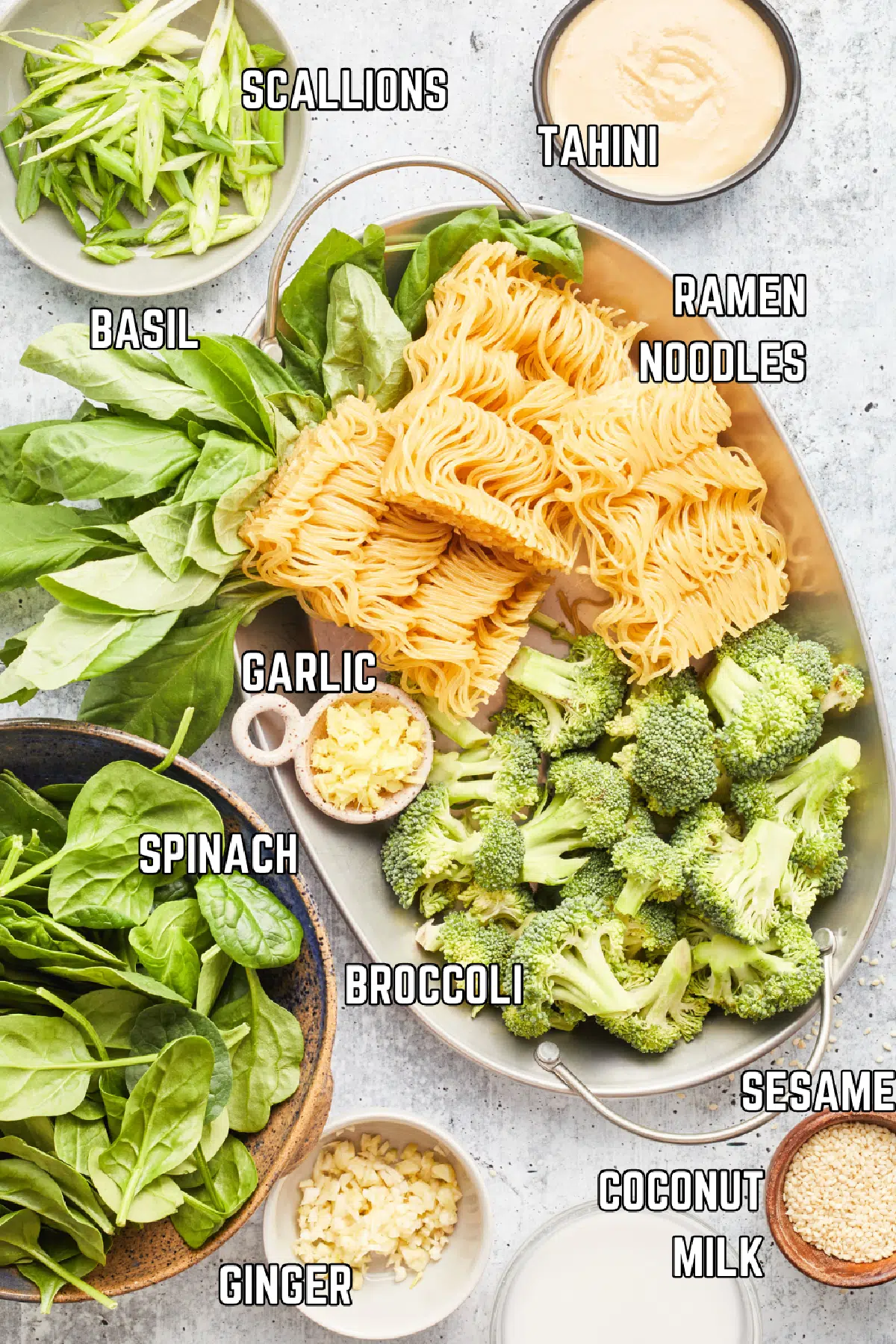 Overhead view of several bowls of ingredients to make ginger sesame noodles: scallions, tahini, fresh basil, ramen noodles, fresh minced garlic, broccoli, spinach, fresh minced ginger, sesame seeds, and coconut milk.