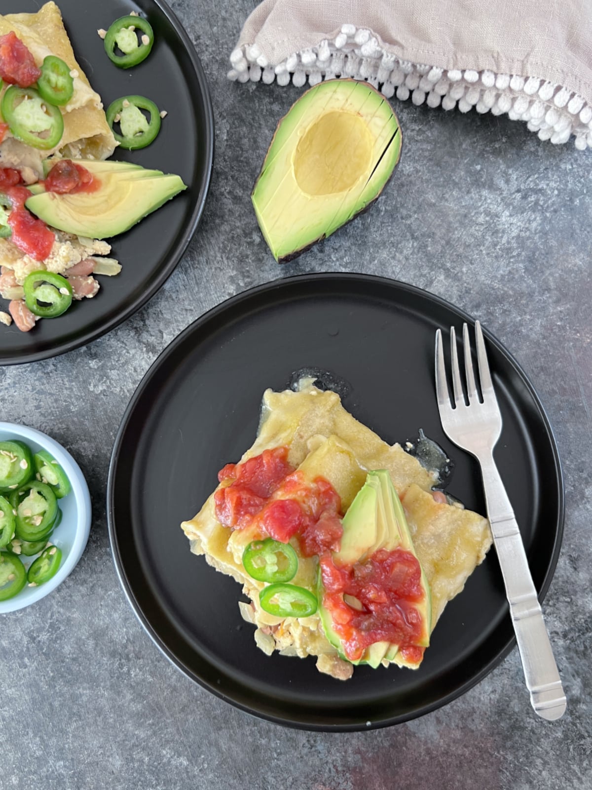 Overhead view of two plates of breakfast enchiladas sit on a dark grey marble surface. Enchiladas are filled with a tofu scramble and beans, and garnished with sliced jalapeños, sliced avocado, and deep red salsa. A half avocado, a small bowl of jalapeno slices, and a tan cloth napkin sit on the side.