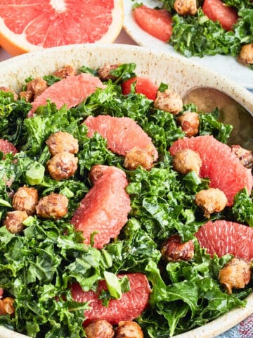 A large stoneware serving bowl of grapefruit kale salad sits on a white surface. Next to the bowl of salad is a fresh grapefruit cut in half, a plate of salad, a few hazelnuts, and a blue cloth napkin.