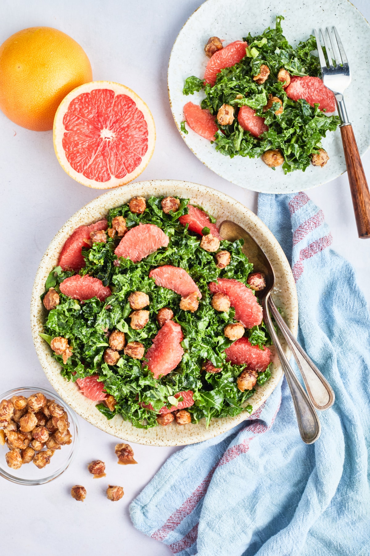 Overhead view of a large stoneware serving bowl of grapefruit kale salad sits on a white surface. Next to the bowl of salad is a fresh grapefruit cut in half, a plate of salad, a few hazelnuts, and a blue cloth napkin.
