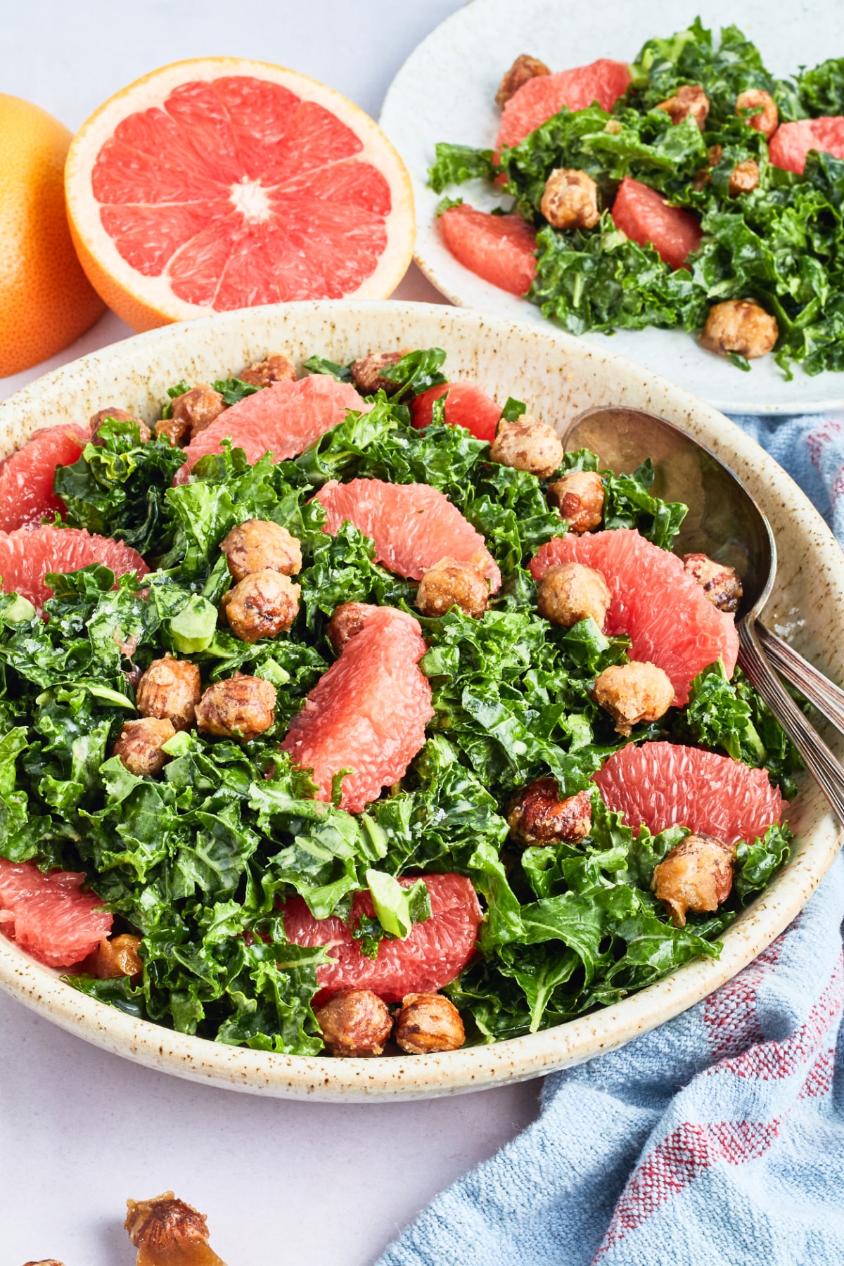 A large stoneware serving bowl of grapefruit kale salad sits on a white surface. Next to the bowl of salad is a fresh grapefruit cut in half, a plate of salad, a few hazelnuts, and a blue cloth napkin.