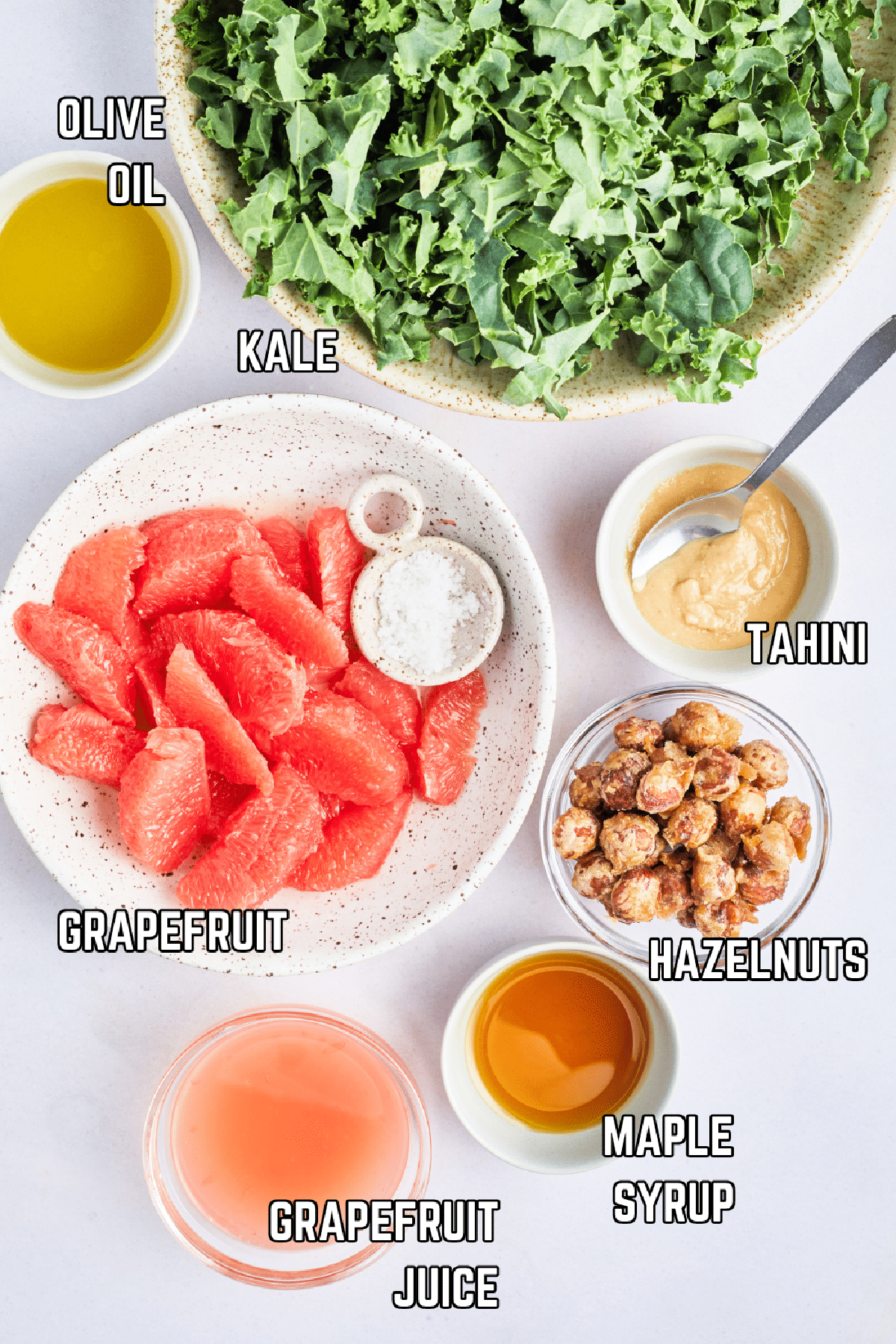 Overhead view of bowls of ingredients to make salad: kale, olive oil, tahini, grapefruit, hazelnuts, maple syrup, and grapefruit juice.