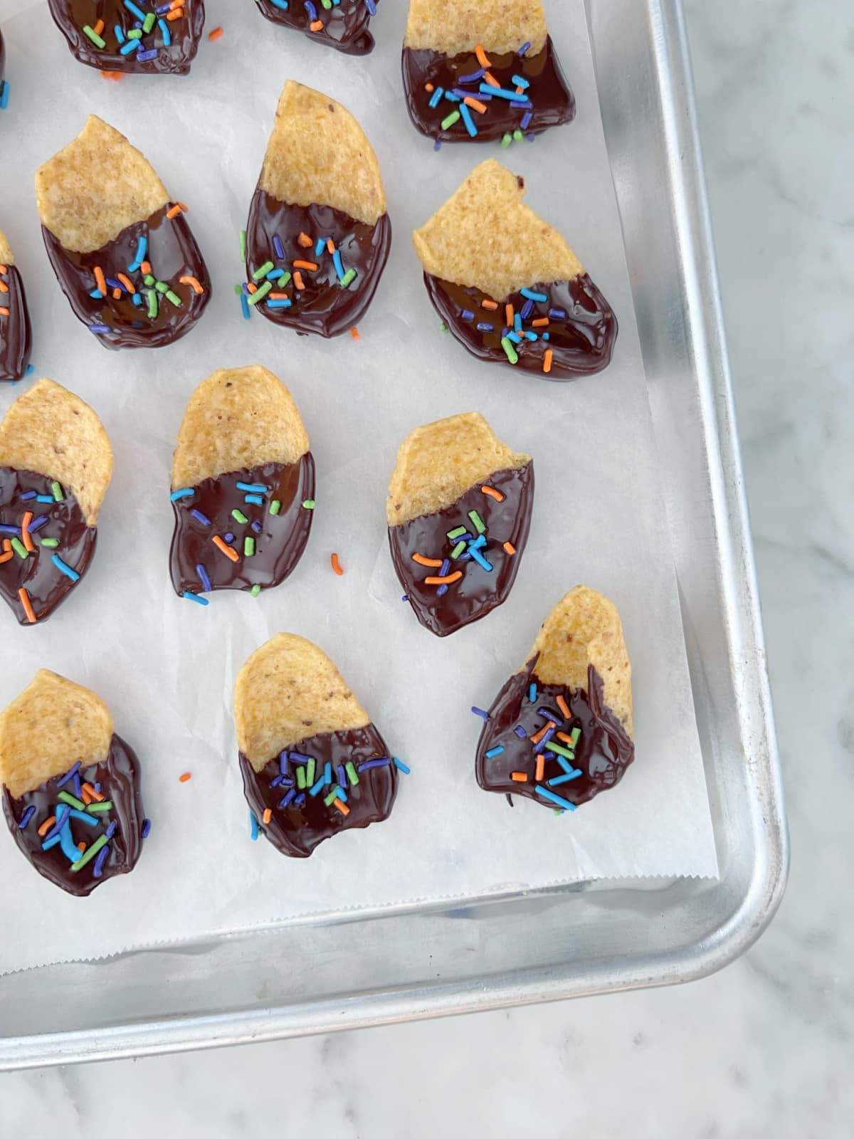 Overhead view of a parchment lined baking pan of chocolate dipped corn chips covered in sprinkles. The chocolate is shiny from being freshly dipped and has not yet dried.