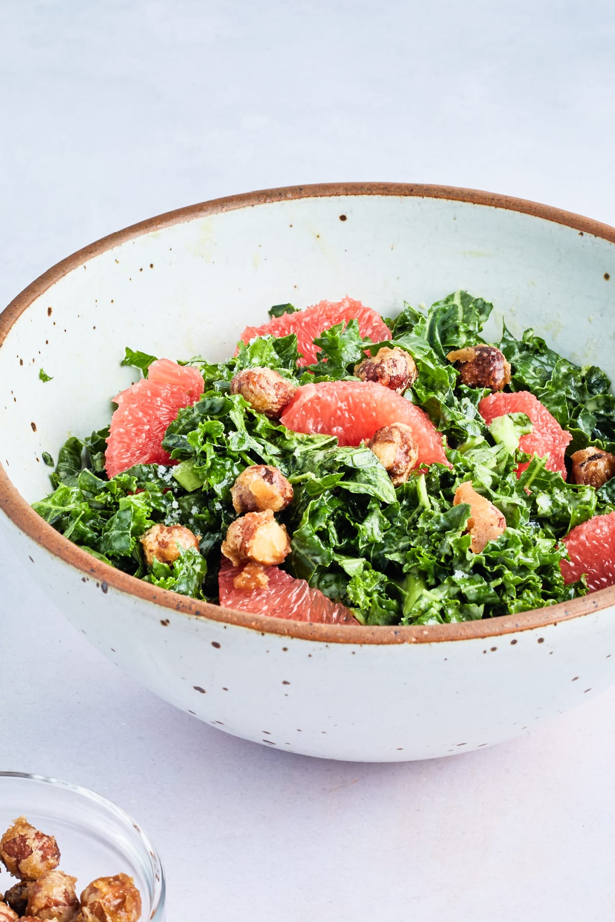 A white rustic stoneware bowl filled with grapefruit kale salad. Salad is thin strips of kale, fresh pink grapefruit segments, grapefruit tahini dressing, and hazelnuts.