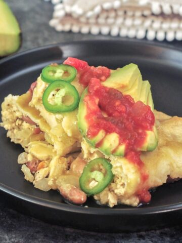One serving of breakfast enchiladas on a matte black plate. Enchiladas are filled with a tofu scramble and beans, and garnished with sliced jalapeños, sliced avocado, and deep red salsa. A half avocado is blurred in the background.
