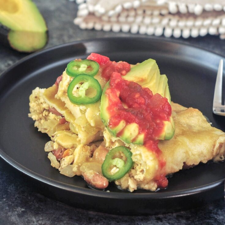 One serving of breakfast enchiladas on a matte black plate. Enchiladas are filled with a tofu scramble and beans, and garnished with sliced jalapeños, sliced avocado, and deep red salsa. A half avocado is blurred in the background.