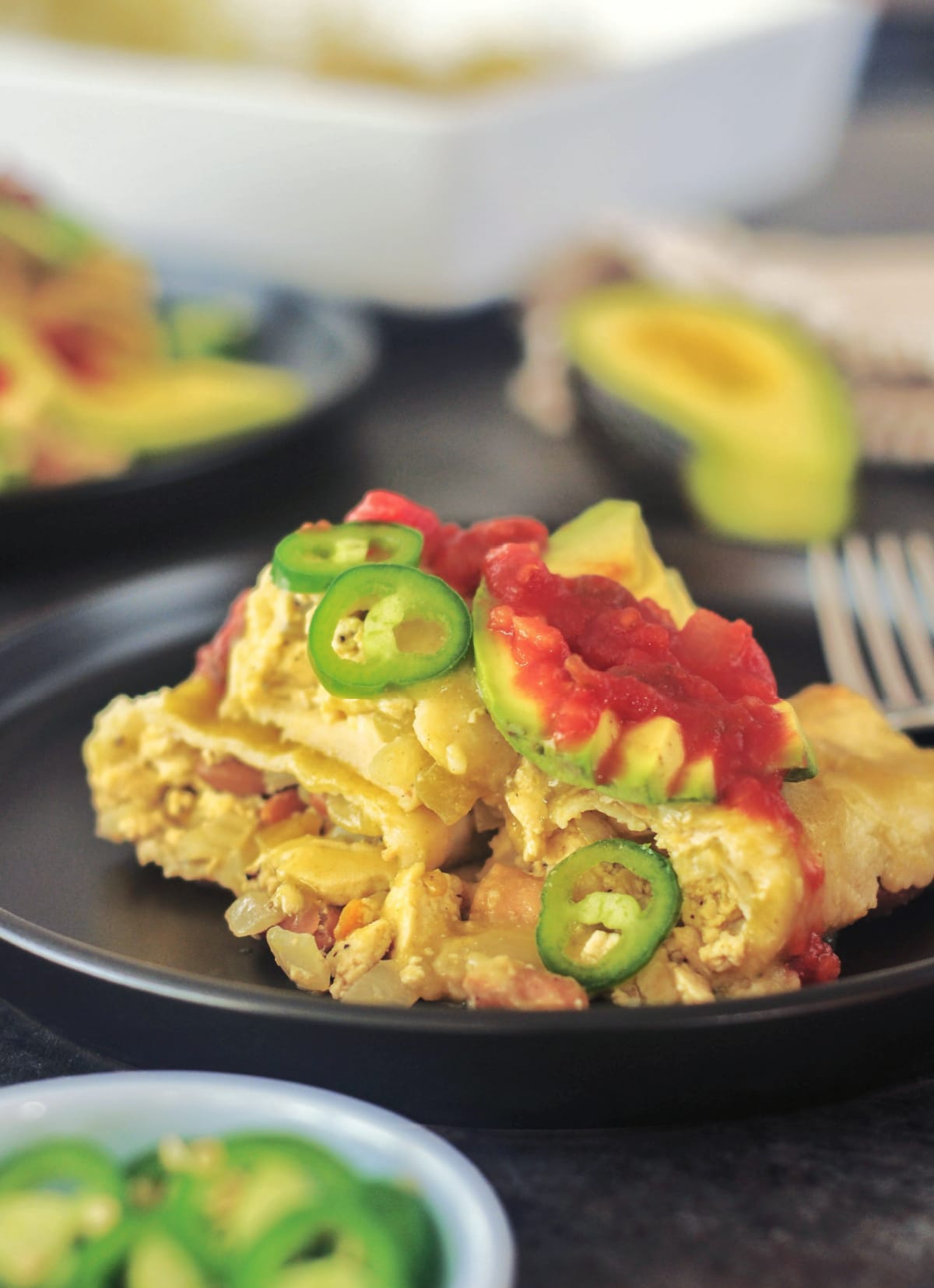 One serving of breakfast enchiladas on a matte black plate. Enchiladas are filled with a tofu scramble and beans, and garnished with sliced jalapeños, sliced avocado, and deep red salsa. A half avocado and the baking dish of enchiladas are blurred in the background.